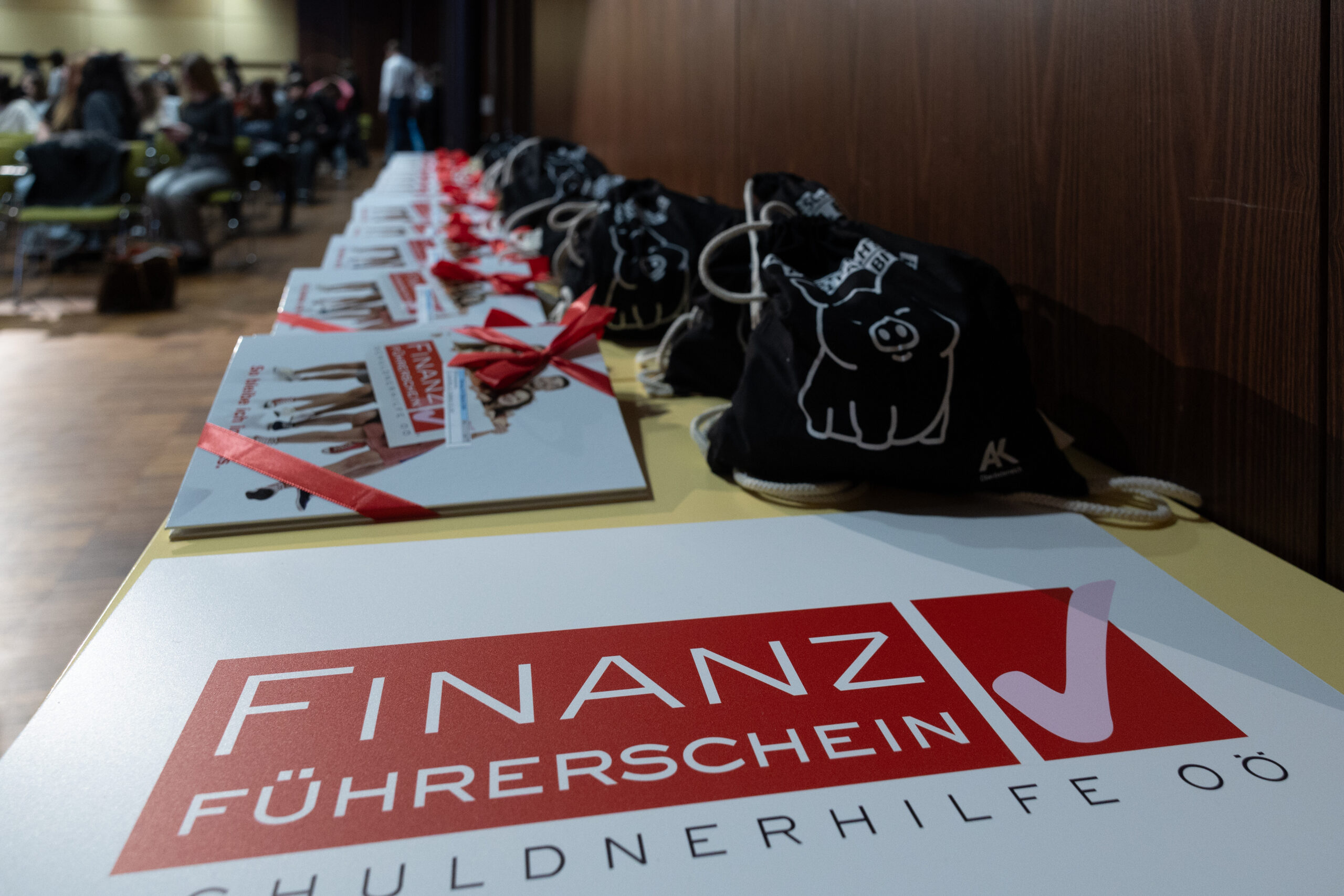 Schild mit Logo OÖ Finanzführerschein - im Hintergrund Mappen mit Zertifikaten und Taschen der AK OÖ Zertifikatsverleihungen OÖ Finanzführerschein