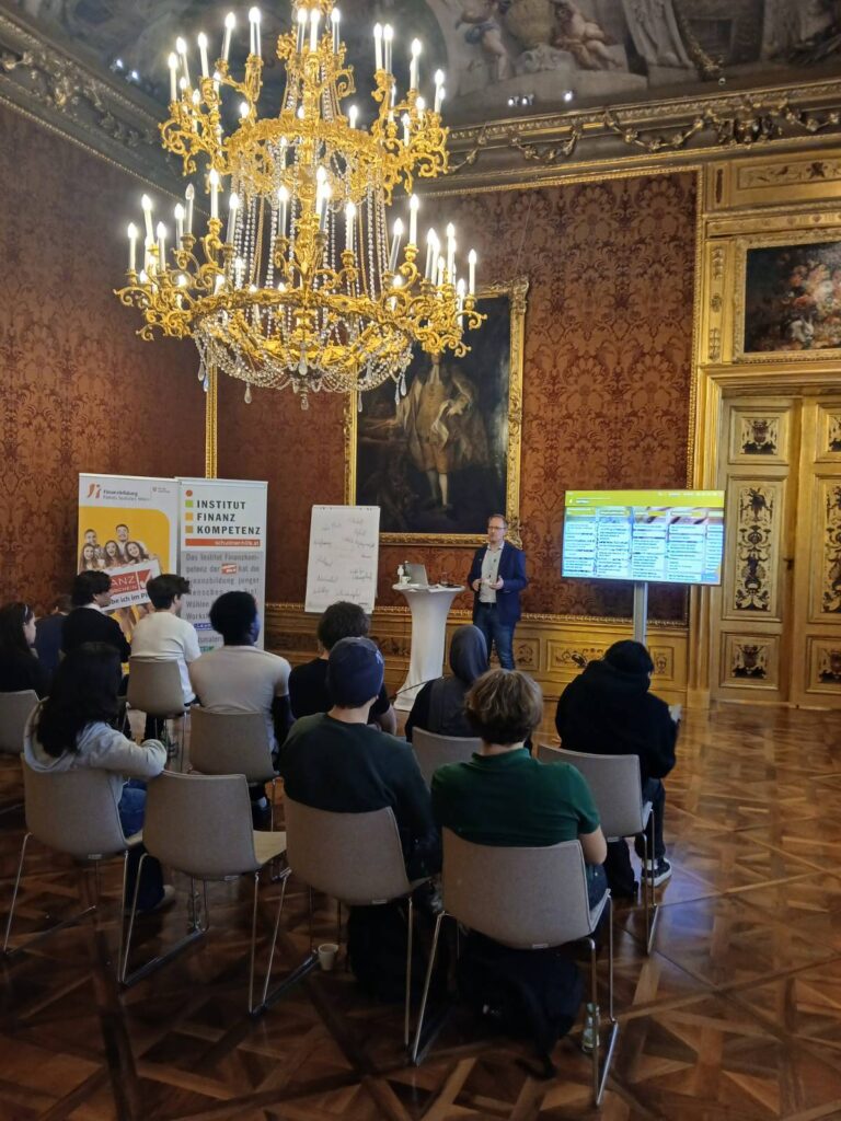 Workshop im Roten Saal des Finanzministeriums. Vortragender im Hintergrund, im Vordergrund unter Kronleuchter sitzend eine Schülergruppe von hinten fotografiert. 